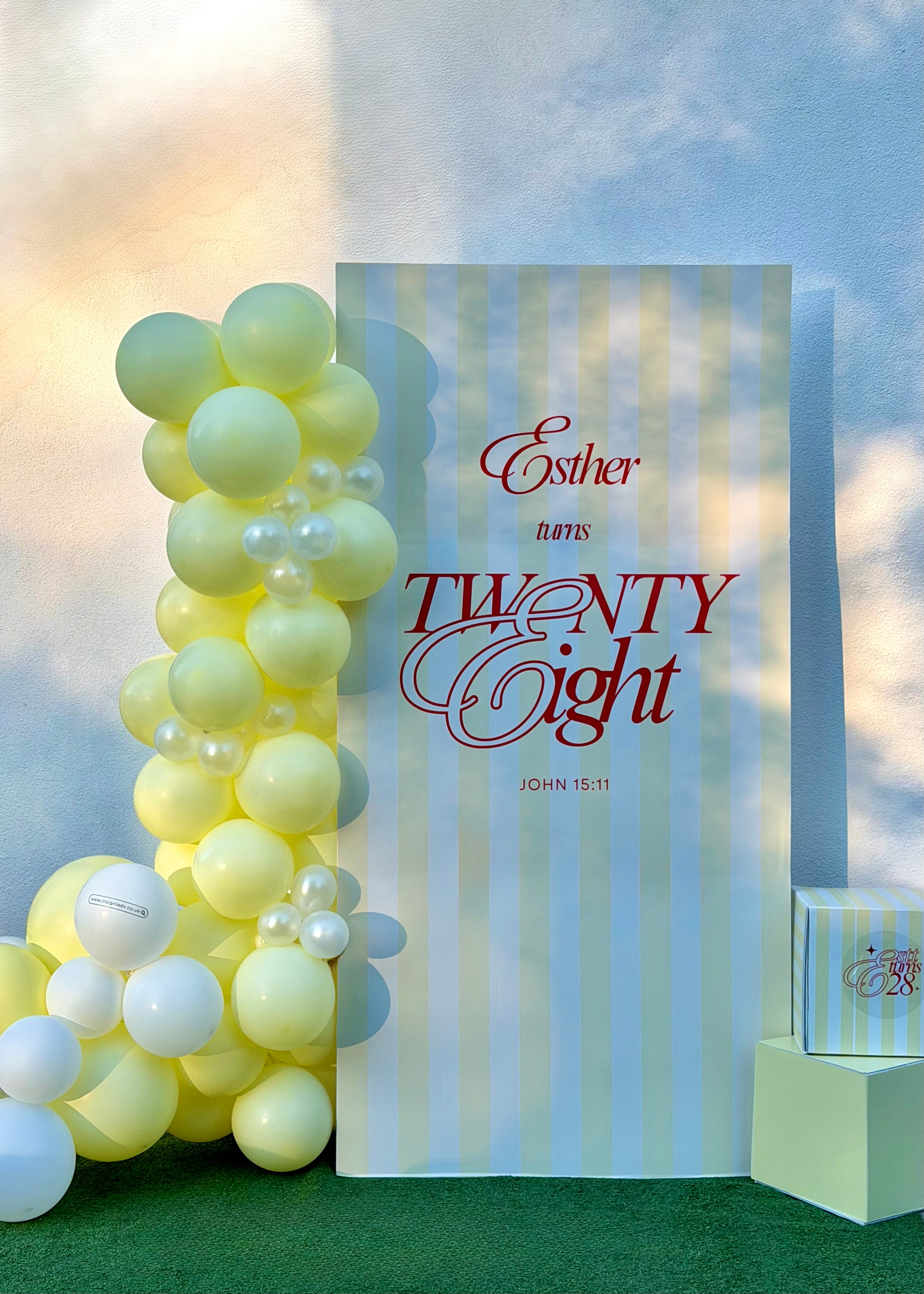 A decorative party backdrop setup featuring a tall white sign with vertical pale blue stripes and red script text reading 'Esther turns TWENTY Eight' with a Bible verse. A cluster of pale yellow and white balloons stands to the left, and a small gift box sits on a pedestal to the right, all set against a softly lit outdoor-style background.