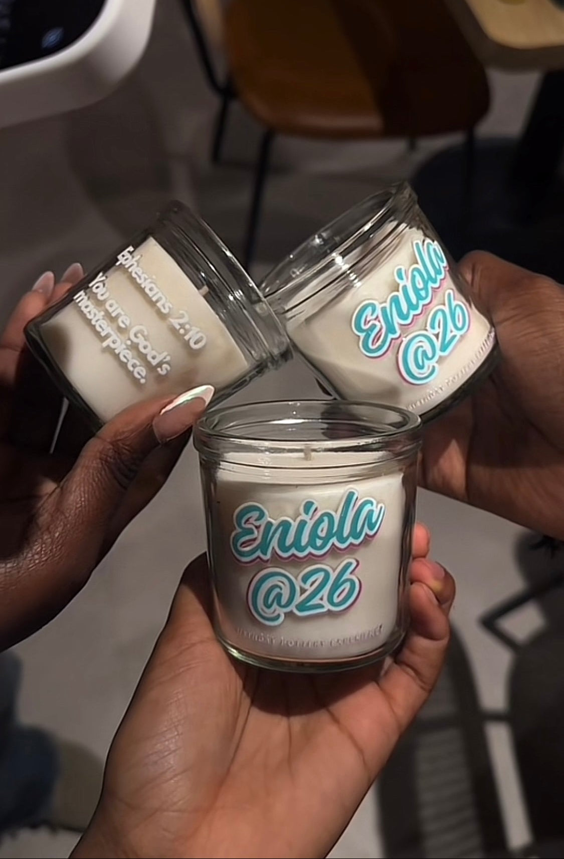 Three clear jars with white candle inside, labeled 'Eniola @26', held by hands against a blurred indoor background.